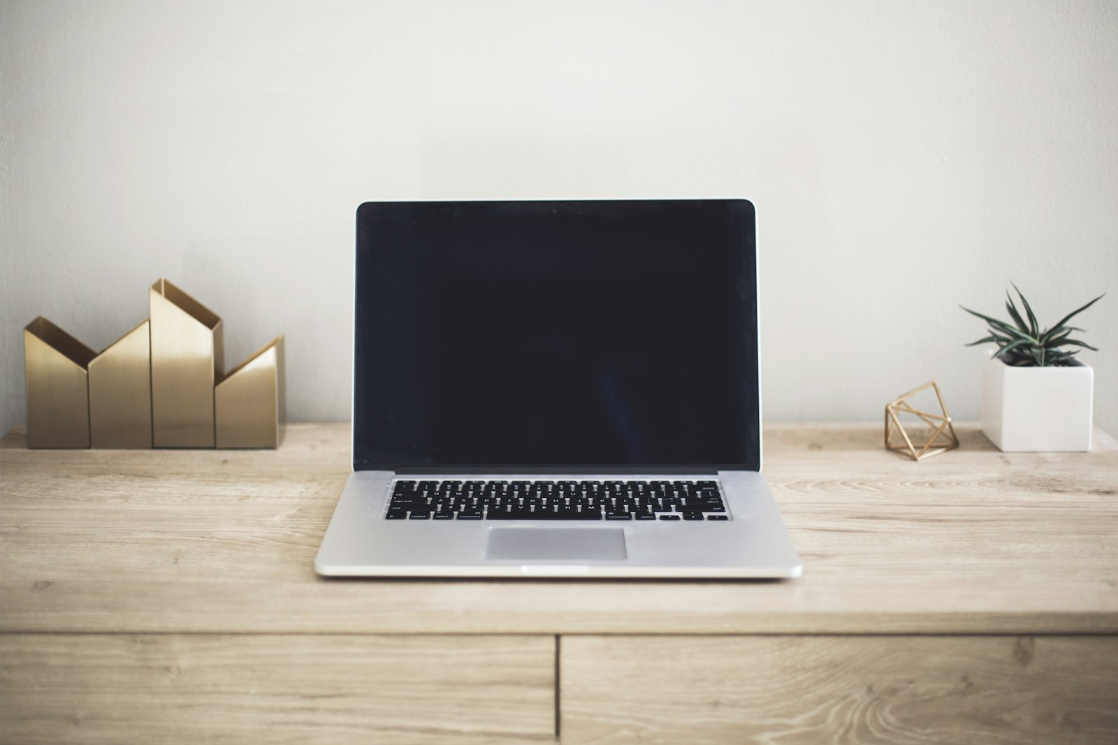 Aufgeräumter, minimalistischer Schreibtisch aus hellem Holz mit einem geschlossenen silbernen Laptop, einer kleinen Pflanze im weißen Topf und goldenen geometrischen Akzenten